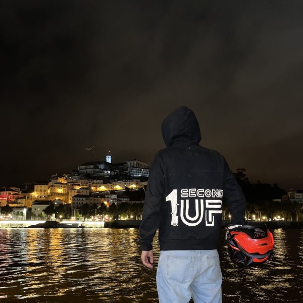Back view of a Second 1UP black motorcycle hoodie with a large logo, worn by a motorsports rider in a helmet. Reflective gear in use